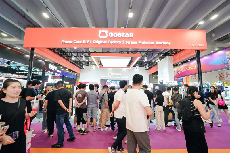 Выставочный стенд GOBEAR, демонстрирующий аппараты для изготовления чехлов для телефонов и оборудование для защиты экрана на торговой выставке, с посетителями, собравшимися вокруг оранжевого стенда